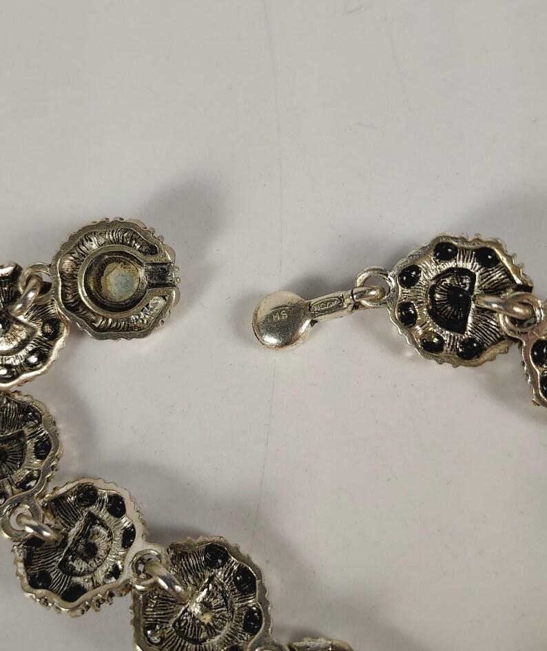 May include: Close-up of a silver necklace with a floral design. The necklace has small, circular links with black accents and a secure clasp. The clasp is connected to a series of decorative links.