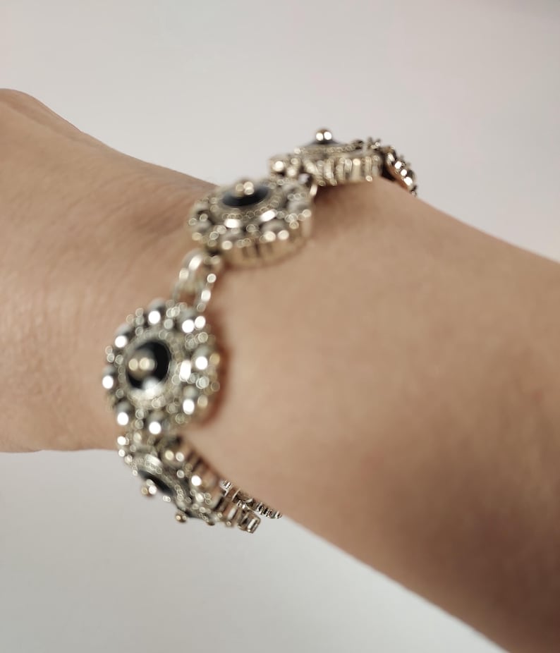 May include: Silver bracelet with a series of decorative circular links. Each link has a black centre and is surrounded by smaller, ornate details. The bracelet is displayed on a person's wrist.