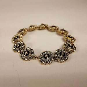 May include: A gold-coloured bracelet featuring a series of circular, flower-like links. Each link has a black centre and is surrounded by small, raised gold beads. The bracelet is linked together with small gold connectors.