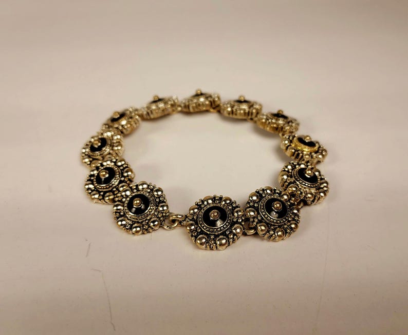 May include: A gold-coloured bracelet featuring a series of circular, flower-like links. Each link has a black centre and is surrounded by small, raised gold beads. The bracelet is linked together with small gold connectors.