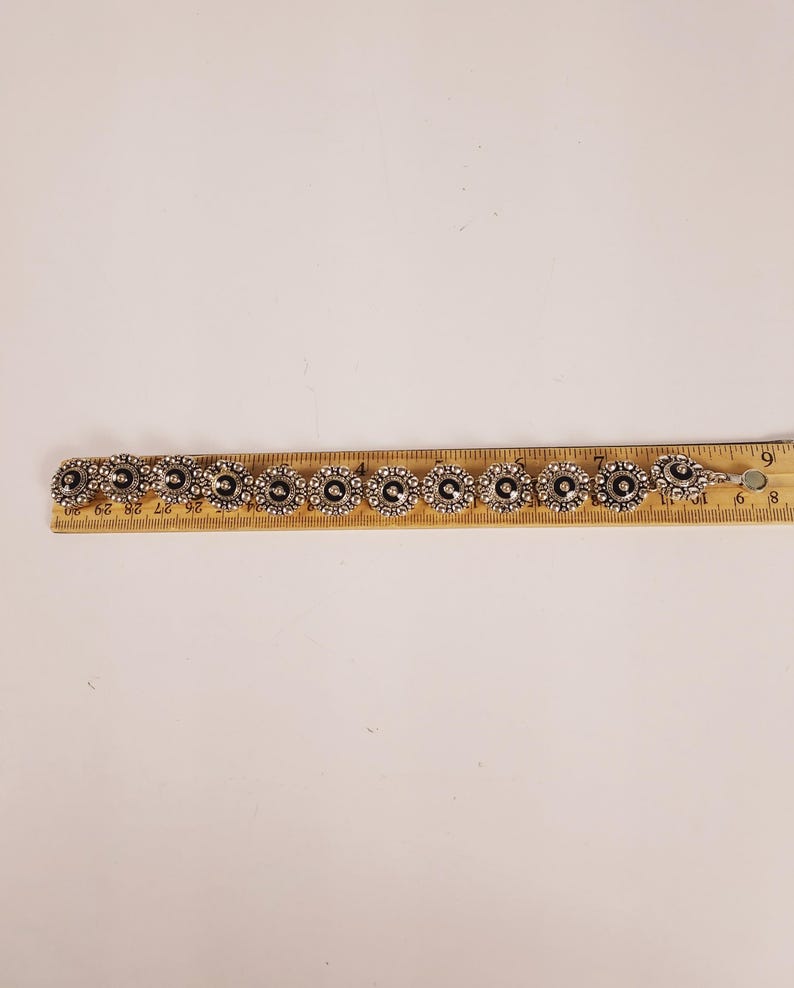 May include: Silver bracelet with circular, flower-shaped links. Each link has a dark centre and a detailed silver border. The bracelet is approximately 17.78 cm long.