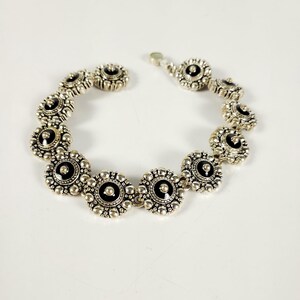 May include: Silver bracelet with a series of flower-shaped links. Each link has a black centre and a detailed, textured border. The bracelet is shown against a white background. The clasp is visible.