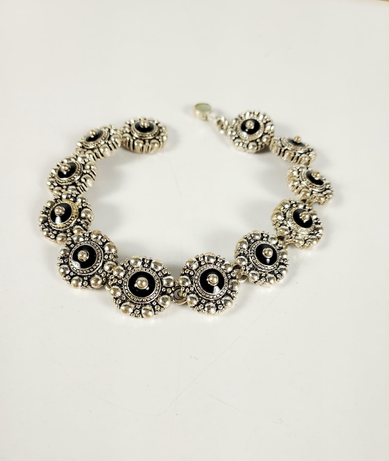 May include: Silver bracelet with a series of flower-shaped links. Each link has a black centre and a detailed, textured border. The bracelet is shown against a white background. The clasp is visible.