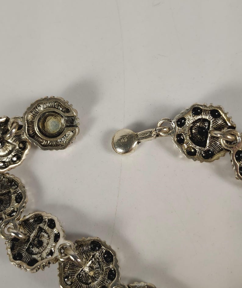 May include: Close-up of a silver necklace with a floral design. The necklace has small, circular links with black accents and a secure clasp. The clasp is connected to a series of decorative links.