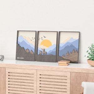 Puede incluir: Tres impresiones artísticas enmarcadas que representan un paisaje de montaña con un sol amarillo, ciervos y pájaros. Las impresiones se exhiben en un mueble de madera clara con una planta y objetos decorativos. La habitación tiene una estética moderna.