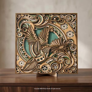 May include: A layered wooden art piece featuring a hummingbird design. The artwork is square, with intricate cut-out details in shades of brown, green, and white. The hummingbird is central, surrounded by floral and geometric patterns. The piece is displayed on a wooden stand.