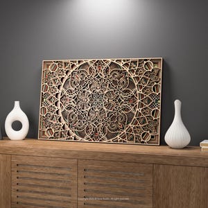 Puede incluir: Una detallada obra de arte de madera en capas con un diseño de mandala se exhibe en un mueble de madera. La obra de arte presenta intrincados patrones recortados en tonos marrones y beige. Dos jarrones blancos y una pequeña pila de libros también están en el mueble.