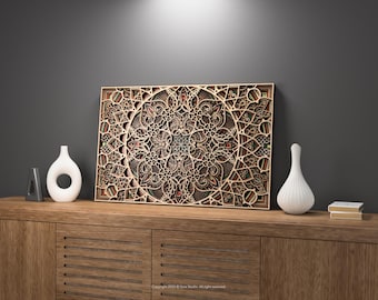 May include: A detailed, layered wooden art piece with a mandala design is displayed on a wooden cabinet. The artwork features intricate cut-out patterns in shades of brown and beige. Two white vases and a small stack of books are also on the cabinet.