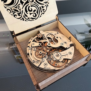May include: A wooden laser-cut watch mechanism replica displayed inside a wooden box. The watch gears and components are intricately detailed. The box lid features a decorative cutout design. The date 26012023 and the number 333 are visible on the watch.