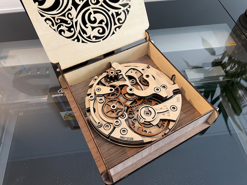 May include: A wooden laser-cut watch mechanism replica displayed inside a wooden box. The watch gears and components are intricately detailed. The box lid features a decorative cutout design. The date 26012023 and the number 333 are visible on the watch.