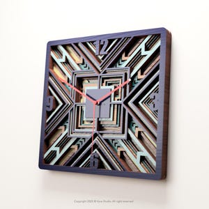 May include: Square wooden wall clock with a layered geometric design. The clock face features a multi-colored pattern with a dark blue frame and red hands. The numbers 12, 9, and 6 are visible.