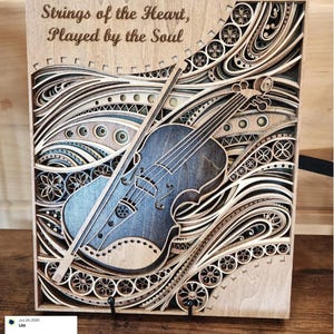 May include: A wooden layered art piece featuring a violin with a bow, set against a backdrop of swirling, decorative patterns. The text "Strings of the Heart, Played by the Soul" is at the top. The artwork is primarily in shades of brown and blue.