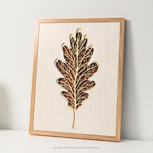 Könnte beinhalten: Gerahmtes Holzkunstwerk mit einem geschichteten Eichenblatt-Design. Das Blatt ist aufwendig geschnitten und zeigt ein detailliertes Muster in Braun- und Goldtönen. Das Kunstwerk ist vor einem hellen Holzgrund, was eine natürliche Ästhetik erzeugt.