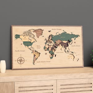 Könnte beinhalten: Eine Weltkarte aus Holz mit einem Naturholzrahmen. Die Karte zeigt Länder in Grün-, Braun- und Beigetönen. Die Ozeane sind beschriftet, und eine Windrose befindet sich unten links. Die Karte wird auf einem Holzschrank präsentiert.