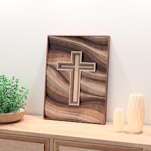 Puede incluir: Una obra de arte de cruz de madera en capas con un acabado de madera natural. La cruz es el punto focal, sobre un fondo de madera ondulada y en capas en varios tonos de marr&oacute;n. La obra de arte est&aacute; enmarcada en un borde marr&oacute;n oscuro.