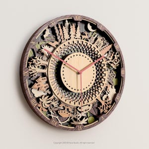 May include: A round wooden wall clock with a layered design featuring trees, houses, and other natural elements. The clock has a light brown face with pink hands and Roman numerals around the edge. The clock is approximately 30 cm in diameter.
