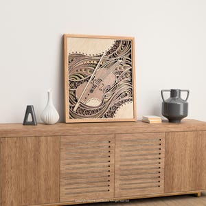 May include: A framed wooden art piece featuring a detailed violin design with a bow. The artwork is in shades of brown and beige, with intricate patterns. The piece is displayed on a wooden cabinet with decorative items.