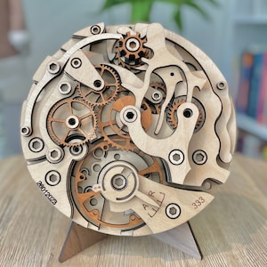 May include: A wooden, layered clock mechanism model with intricate gears and details. The model is made of light-colored wood with darker wood accents. The date 20012023 and the number 333 are visible on the face. It is displayed on a wooden stand.