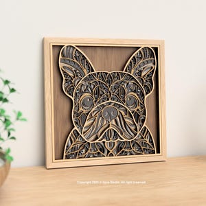 Puede incluir: Una obra de arte de madera en capas que representa la cara de un bulldog francés. Los rasgos del perro están intrincadamente cortados de varias tonalidades de madera gris y marrón, enmarcados en un marco de madera natural. La obra se exhibe sobre una superficie de madera.