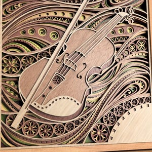 May include: A layered wooden art piece featuring a violin and bow. The artwork uses various shades of brown, green, and yellow, with intricate patterns and designs. The violin is the central focus, with the bow resting across it. The piece is framed in a light-colored wooden frame.