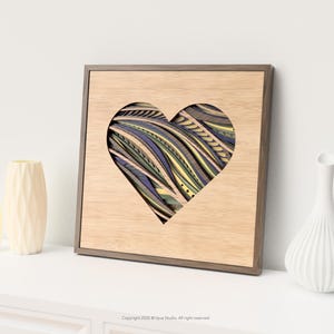 Può includere: Opera d'arte da parete in legno con un ritaglio a forma di cuore che rivela un design astratto colorato a strati. La cornice è in legno naturale. Due vasi decorativi sono a sinistra e un vaso bianco a destra.