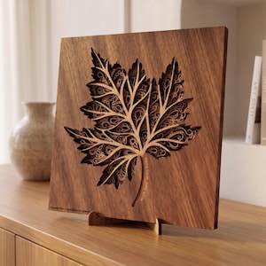 Könnte beinhalten: Quadratische Wandkunst aus Holz mit einem detaillierten Ahornblatt-Design. Das Blatt ist aufwendig mit einem geschichteten Effekt geschnitzt, der ein hellbraunes Inneres vor einem dunkleren braunen Hintergrund zeigt. Das Kunstwerk wird auf einem Holzständer präsentiert.