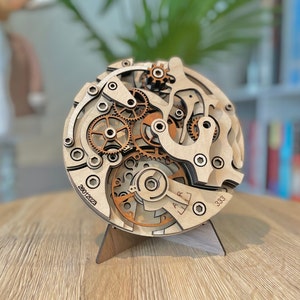 May include: A wooden clock mechanism model with intricate gears and components, displayed on a wooden stand. The model features a layered design with visible gears in shades of brown and natural wood. The numbers "28012023" and "333" are visible.