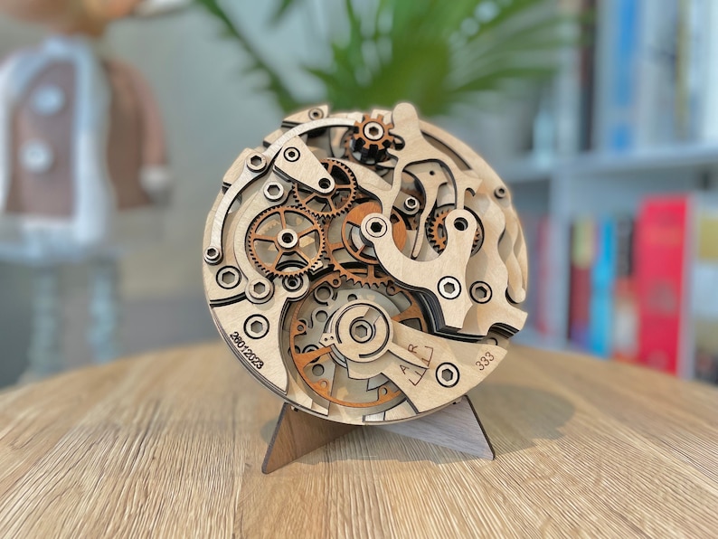 May include: A wooden clock mechanism model with intricate gears and components, displayed on a wooden stand. The model features a layered design with visible gears in shades of brown and natural wood. The numbers "28012023" and "333" are visible.