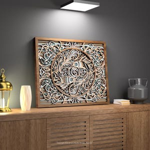 Könnte beinhalten: Ein mehrschichtiges Wandkunstwerk aus Holz mit einem aufwendigen, kreisförmigen Design. Das Kunstwerk hat einen braunen Rahmen und ein detailliertes Muster in Weiß, Braun und Türkis. Es wird auf einem Holzschrank zusammen mit einer goldenen Laterne und einer grauen Vase ausgestellt.