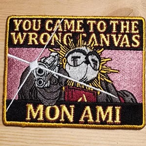 May include: Embroidered patch with the text "YOU CAME TO THE WRONG CANVAS MON AMI". The patch features a cartoon character with sunglasses holding a gun, set against a pink and black background with gold trim.