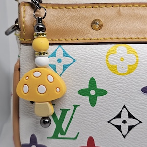May include: A yellow mushroom charm with white spots and a beaded accent hangs from a silver clasp. The charm is attached to a white purse with colorful geometric patterns and brown trim.