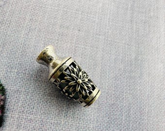 Könnte beinhalten: Eine kleine, silberfarbene, vasenförmige Perle mit floralem Design. Die Perle hat ein detailliertes, durchbrochenes Muster um den Körper und eine strukturierte Oberfläche. Die Perle ist wahrscheinlich für die Schmuckherstellung oder andere Bastelarbeiten gedacht.