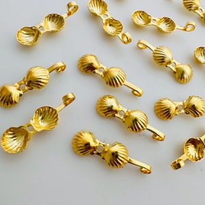 May include: A group of gold-colored metal shell-shaped charms with a loop at the top. The charms are arranged on a white surface.