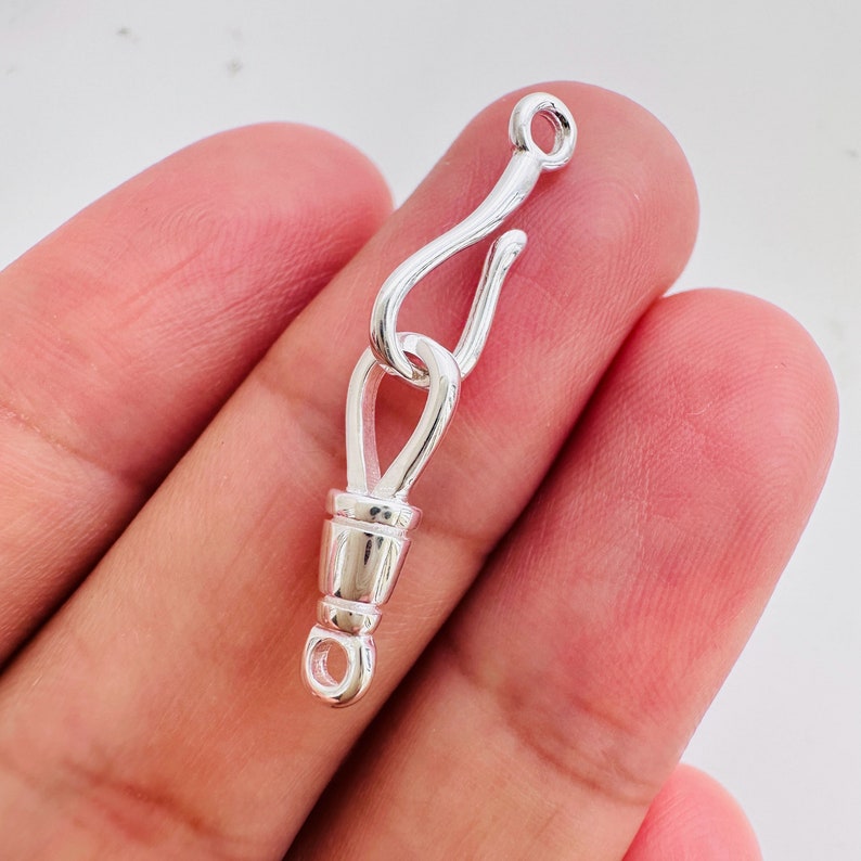 May include: Silver-toned metal clasp with a hook and a barrel-shaped closure. The clasp is designed for attaching to a chain or cord.
