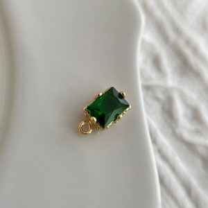 May include: Gold charm with a green emerald-shaped gemstone. The charm has a small loop for attaching to a necklace or bracelet.