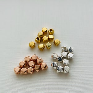 May include: A collection of gold, silver, and rose gold metal beads. The beads are shaped like small, faceted pyramids.
