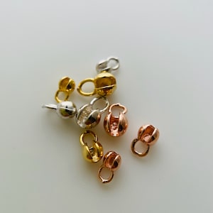 May include: A collection of six gold, silver, and rose gold metal clasps. The clasps are round and have a small loop on one side. They are used for attaching jewelry or other items.