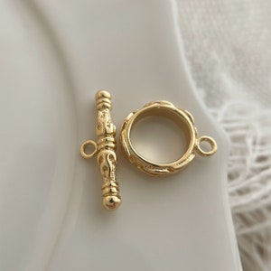 May include: Gold-toned metal toggle clasp with an ornate design. The clasp is made of two pieces: a round ring and a bar with a loop on one end.