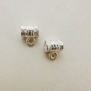 May include: Two silver metal barrel-shaped bead caps with a decorative design and a small loop at the bottom.