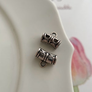 Könnte beinhalten: Zwei kleine, silberfarbene Metall-Schmuckkomponenten. Jede Komponente ist zylindrisch mit einem dekorativen Muster und einer kleinen Schlaufe an einem Ende. Sie werden wahrscheinlich für die Schmuckherstellung verwendet, wie z. B. für Armbänder oder Halsketten.