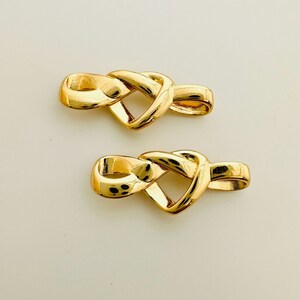 May include: Two gold-toned metal jewelry clasps with an intricate knot design. The clasps are shaped like a heart with a loop on each end.
