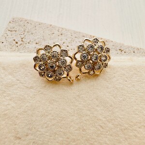 Könnte beinhalten: Ein Paar goldfarbene, blumenförmige Verschlüsse aus Metall mit klaren Strasssteinen. Die Verschlüsse haben eine Schlaufe an der Seite, um sie an einer Halskette oder einem Armband zu befestigen.