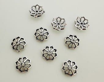 May include: A collection of nine silver-colored, flower-shaped bead caps. Each cap features a central hole surrounded by a series of petals and decorative cutouts. The caps are scattered on a light-colored surface, showcasing their intricate design.