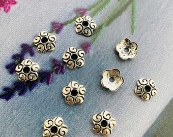 May include: A collection of silver-colored, flower-shaped beads with a central hole and swirling design. The beads are scattered on a light blue surface with a floral pattern in the background. These decorative beads are ideal for jewelry making.