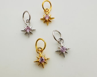 May include: Four starburst charms, two in gold and two in silver, each with a small pink gemstone center. The charms are designed to be attached to jewelry. The background is a solid, light color.