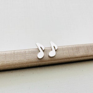 Könnte beinhalten: Ein Paar silberfarbene Ohrstecker in Form von Noten. Die Ohrringe sind klein und haben ein schlichtes Design. Sie werden auf einer strukturierten, metallischen Oberfläche präsentiert.