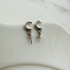 May include: A pair of silver earrings. Each earring has a curved, C-shaped top with a small, dangling post and a round, flat base. The earrings are presented on a white surface.