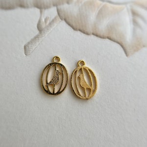May include: Two gold-toned metal charms in the shape of a bird inside a cage. The charms are oval-shaped and have a loop at the top for attaching to a necklace or bracelet.