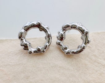 May include: A pair of silver-colored, organic-shaped stud earrings. Each earring is a ring with an irregular, textured surface, featuring small, rounded protrusions. The earrings are displayed on a light beige fabric.