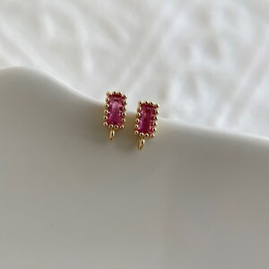 Op de afbeelding: Een paar gouden oorbellen met roze rechthoekige edelstenen. De oorbellen hebben een parelrand rond de edelstenen.